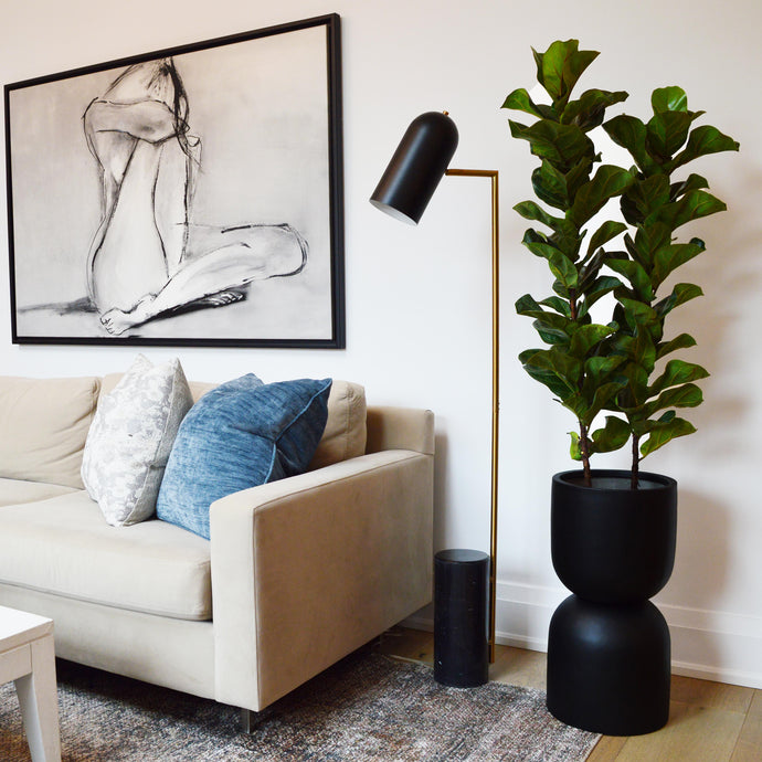 A black hourglass-shaped double stacked pot planter placed on a floor next to a white sofa with a plant inside, in a room with a painting on the wall and a lamp.