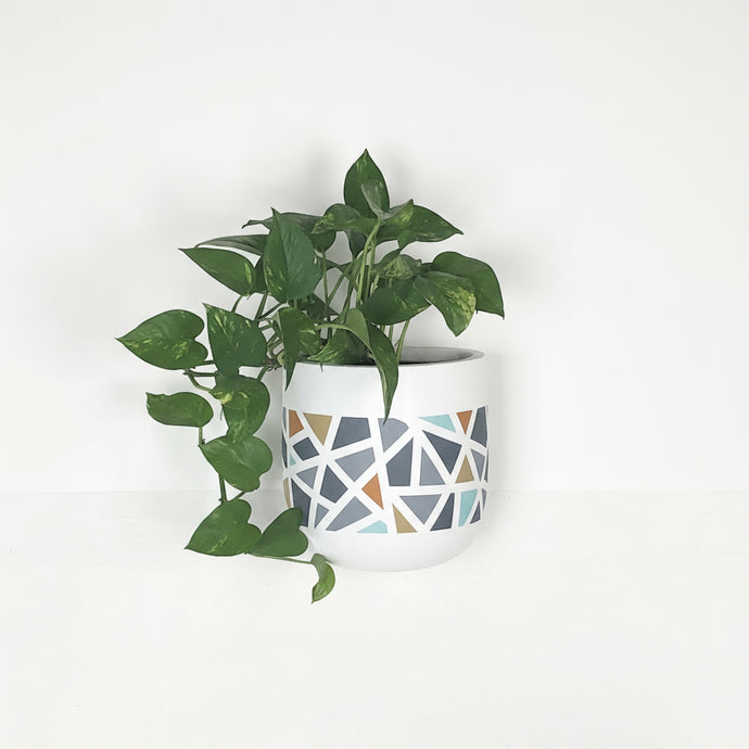 Green potted plant in a white hanging planter pot with geometric patterns on a light gray background