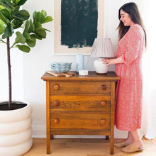 Woman in a red dress holding a lamp next to a wooden dresser and large potted plant.