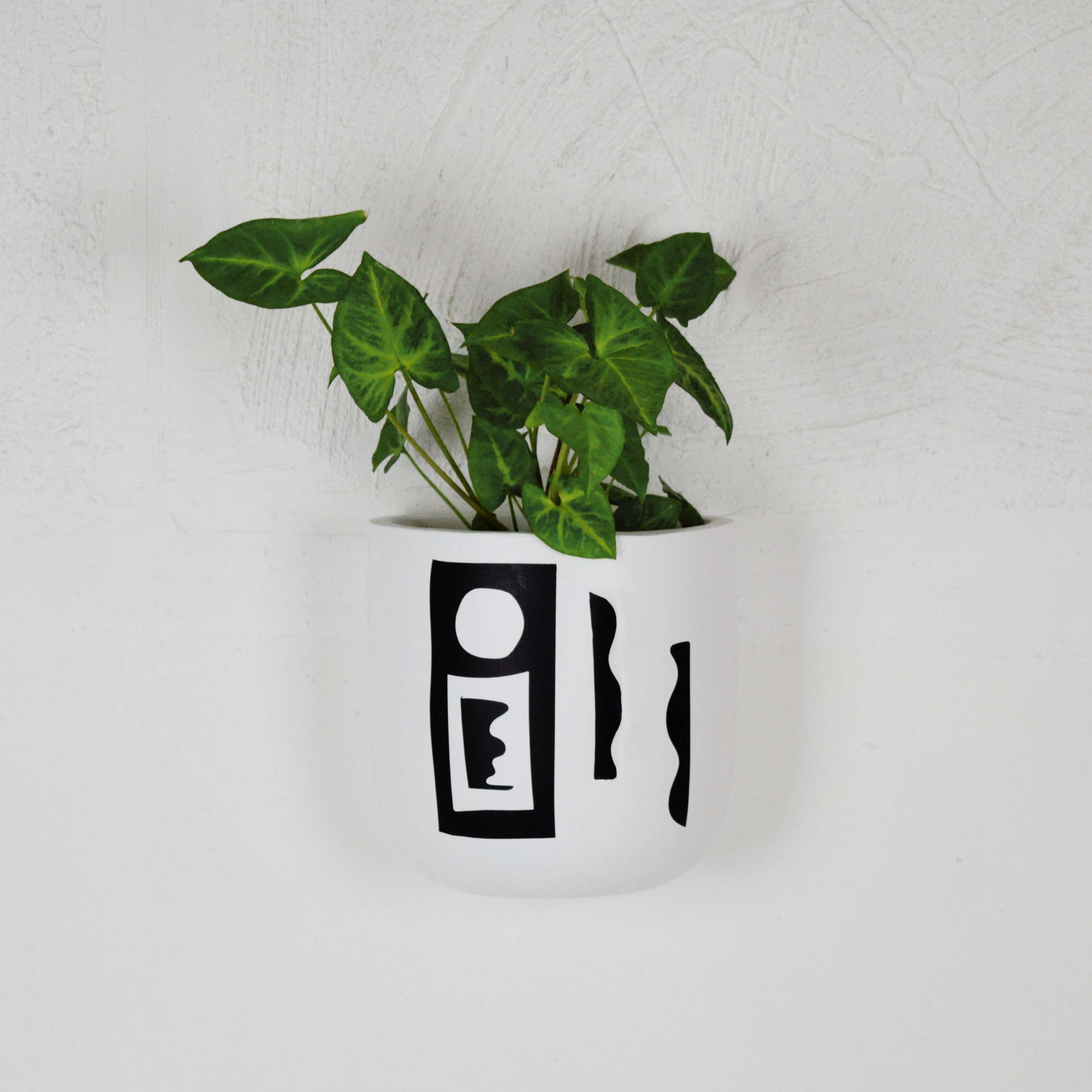 A white, rectangular wall planter with a geometric pattern on the front, mounted on a wall with a green plant inside it.