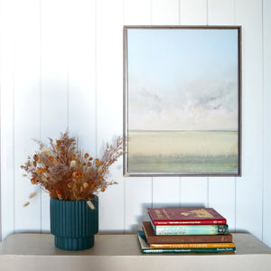 stacked books sit underneath a framed scenic painting.  A small table top scallop pot hold an array of fall inspired dried florals