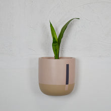 A hand painted beige and clay colored wall planter with a plant growing out of it, attached to a wall.
