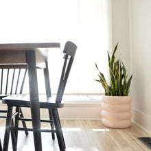 Soft pink stacked-look planter pot in corner of dining room.  A rustic dark wood table with black wooden dining chairs is off to the left.  Soft sunlight warms the room in subtle shades.