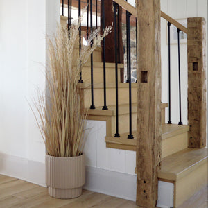 rustic staircase with black metal spindles with a beige fluted plant pot at base.  Tall, dried wheatgrass stands in the plant pot.  Off to the left, there is a framed art print.  A corner of a white kitchen island with black hardware is on the left.  Minimalist plant pot perfect for new and mature plants and florals. Hand painted, lightweight and modern.