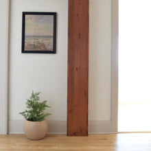 light borwn round sphere plant pot sits on floor with a small tree.  A large walnut support beam is centered in the room and a black framed scenic print hangs above the planter pot.