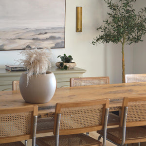 A dining room with cane side chairs and a large dining table has a nude coloured plant pot on the table top with pampas grass.  A sage sideboard can be seen in the background with a large print hanging above it and hanging gold candle sconces.  An artificial olive tree sits in the corner. 