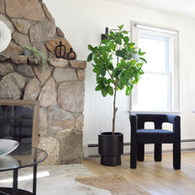 Large stone work fireplace with decorative mantle pieces.  A black pedestal planter sits beside fireplace with a ficus tree.  A stature black arm chair is placed beside it.  A jute rug lays underneath a cow hide rug.  An open window lets in a lot of natural light.