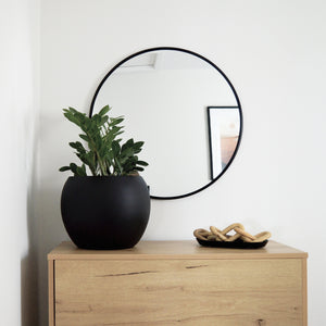 Round black planter pot on top of an oak dresser with a large black round mirror hanging on the wall.  A small black dish holds wooden links.