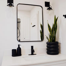 black ring stacked planter with snake plant on corner of bathroom vanity.  Black faucets, black mirror and black sconce lights decorate this bright white bathroom.