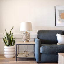 Modern charcoal sofa with framed abstract art print hung above it.  A black metal and oak accent table has a wicker woven lamp on it, with gold accents.  A stacked, bubble planter sits on the floor with a sanservia plant in it.
