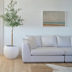 Living room with cream coloured sofa.  Above sofa hangs a print of a beach scene in a white frame.  Beside the couch a large, white sphere planter has a tall olive tree.  A jute rug is layered by a cow hide rug on the floor.