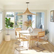A white, round, stacked-look planter with deep channels, placed on a floor beside a chair and a window, with a plant inside it.