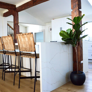 large kitchen island with woven leather bar chairs.  Large walnut support beams line the ceiling and a small black bowl planter sits on floor with an artificial fiddle leaf fig tree