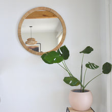 a round rattan mirror hangs on a white wall. A monstera plant is placed on a stool in the corner, in a round blush pink flower pot