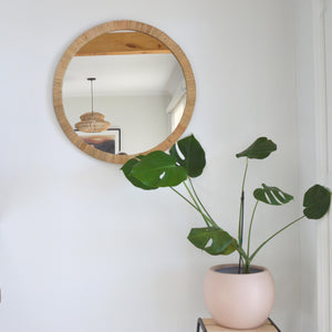 a round rattan mirror hangs on a white wall. A monstera plant is placed on a stool in the corner, in a round blush pink flower pot