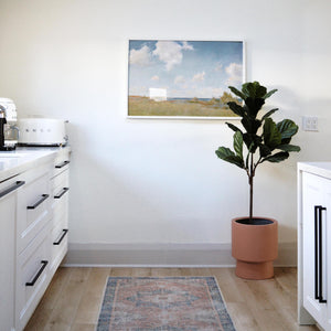 bright white kitchen with black hardware.  Smeg appliances on countertop, a vintage oriental runner is centered on the floor and a pedestal planter with fiddle leaf fig tree is placed off to the right side.  Scenic art print with white frame hangs on the wall