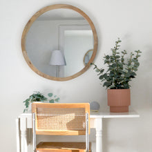 rattan side chair placed at white collapsable desk.  Google home and a small terracotta planter with eucalyptus is on top of desk.  A round oak framed mirror hangs above the desk.