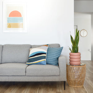 Heather grey couch with colourful cushions and a colourful modern art print hanging above the sofa.  A small terracotta coloured bubble tier planter pot sits on a rattan side table with a tall sanservia snake plant.  A small round mirror hangs on the wall in the distance.