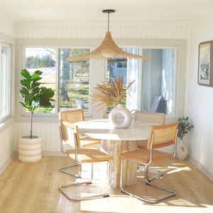 Four cane side chairs placed around a round marble table.  A rattan hanging pendant hangs above table.  An H&M donut vase is placed on centre of table with natural dried palm leaves.  A cream coloured, tiered, ring stacked planter pot sits in the corner holding a fiddle leaf fig plant.  Warm sunlight fills the room from the left side. Plants are powerful.