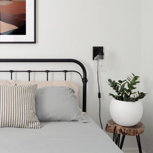 A bed with a black metal bed frame is placed against a white wall.  A black sconce with an eddison bulb is placed to the side.  A small, white bowl shaped planter sits on a wooden night table with a ZZ plant.  A black framed art print hangs above the bed.